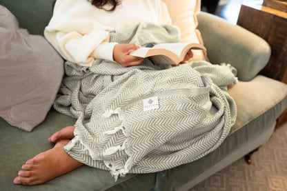 An image of a woman snuggled under the soft cotton throw blanket while reading.